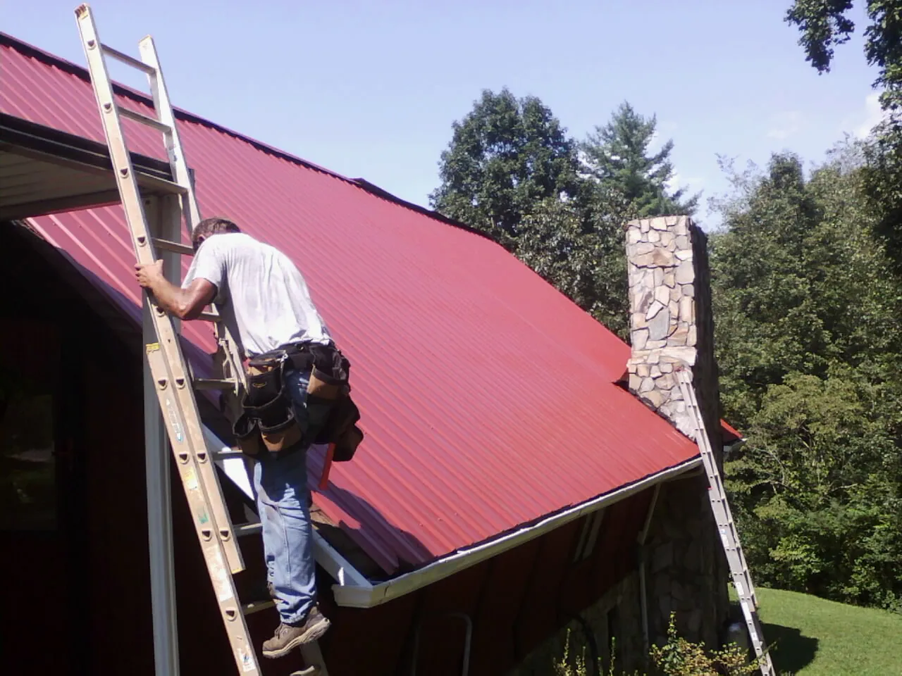 Roofer on a ladder beside a red metal roof and stone chimney — Soffit & Fascia Repair by Roofing Brothers in Sale Creek